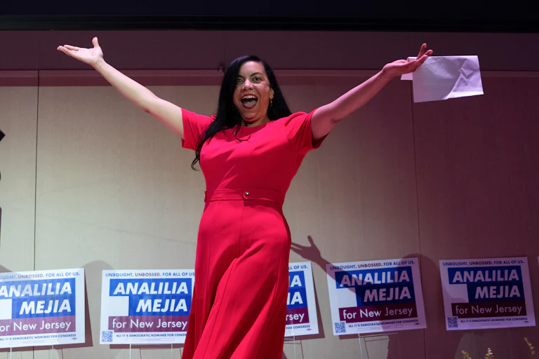 Analilia Mejia raises her arms and smiles with her mouth open while walking on stage. She holds a piece of paper in one hand.