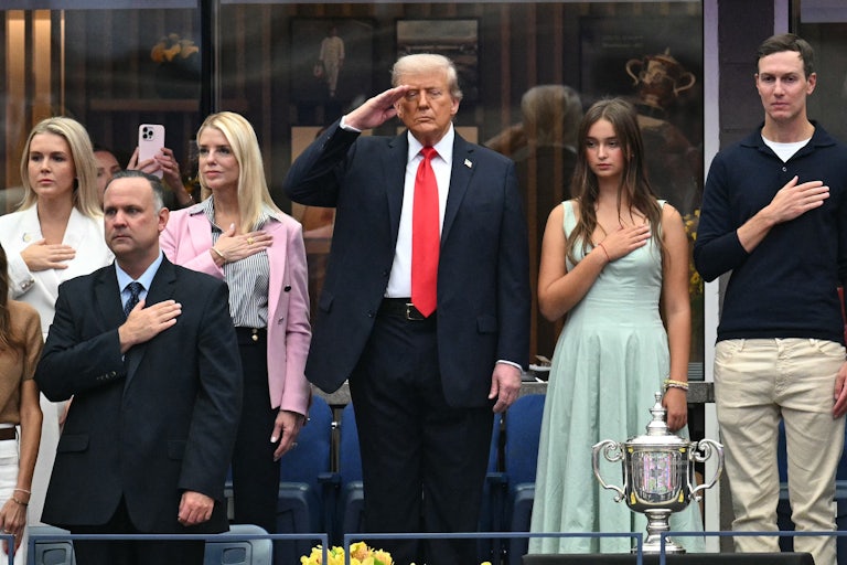 Donald Trump salutes, as Karoline Leavitt, Pam Bondi, Arabella Kushner, and Jared Kushner stand near him with their hands on their heart.