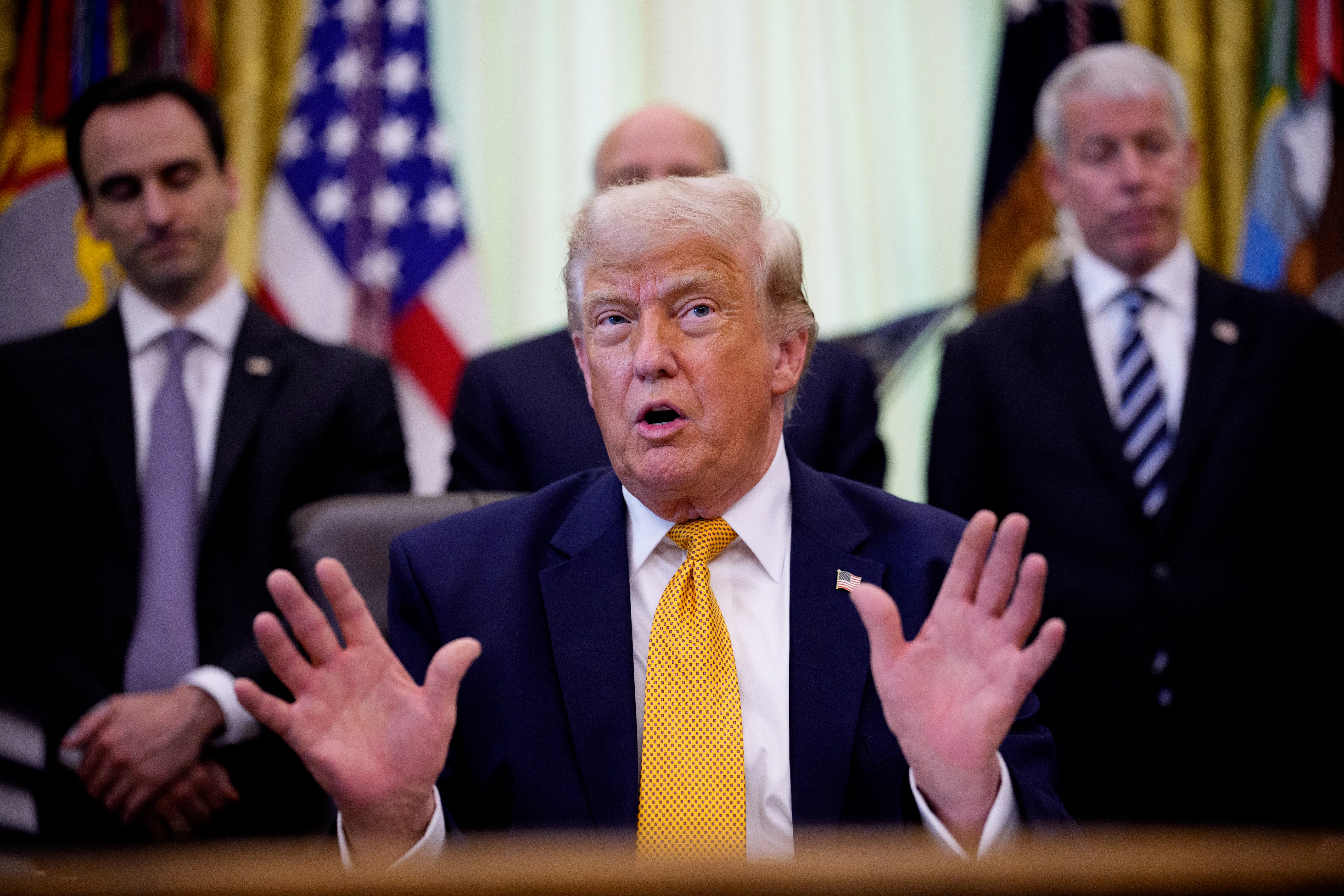 Donald Trump speaking at his desk in the Oval Office of the White House, while others stand behind him.