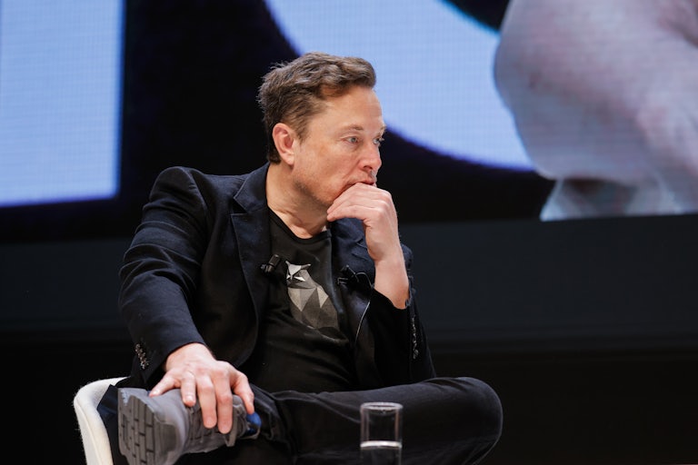 Elon Musk sits in a chair and rests his chin on his hand, looking off into space