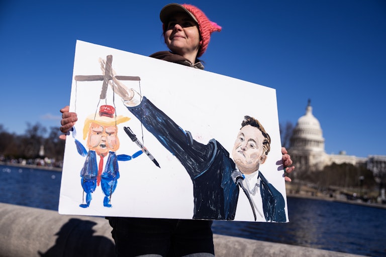 A person stands by the U.S. Capitol and holds up a drawing of Elon Musk doing a Roman salute and using his outstretched hand to control a puppet of Donald Trump