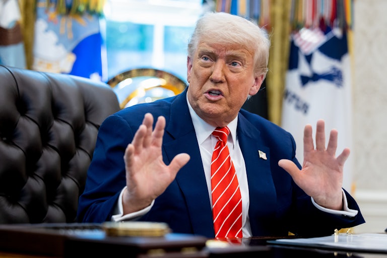 Donald Trump sits at his desk in the Oval Office of the White House and raises his hands as if in defense while speaking.