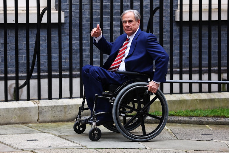 Greg Abbott rolls his wheelchair forward on the sidewalk and makes a thumbs up sign. A black fence is behind him.