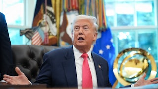 Donald Trump holds his hands out to the side and speaks while sitting at his desk in the Oval Office