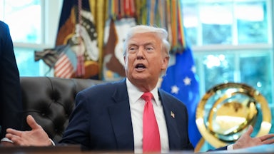 Donald Trump holds his hands out to the side and speaks while sitting at his desk in the Oval Office