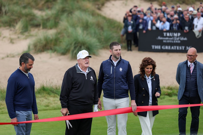 Donald Trump holds a giant pair of scissors and speaks while standing with his adult sons in front of a red ribbon, during the opening of a new golf club