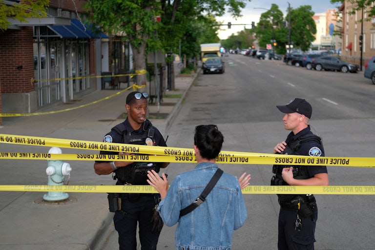 Two police officers stand at a scene with yellow caution tape that says "POLICE DO NOT CROSS." A woman speaks to them on the other side of the tape.