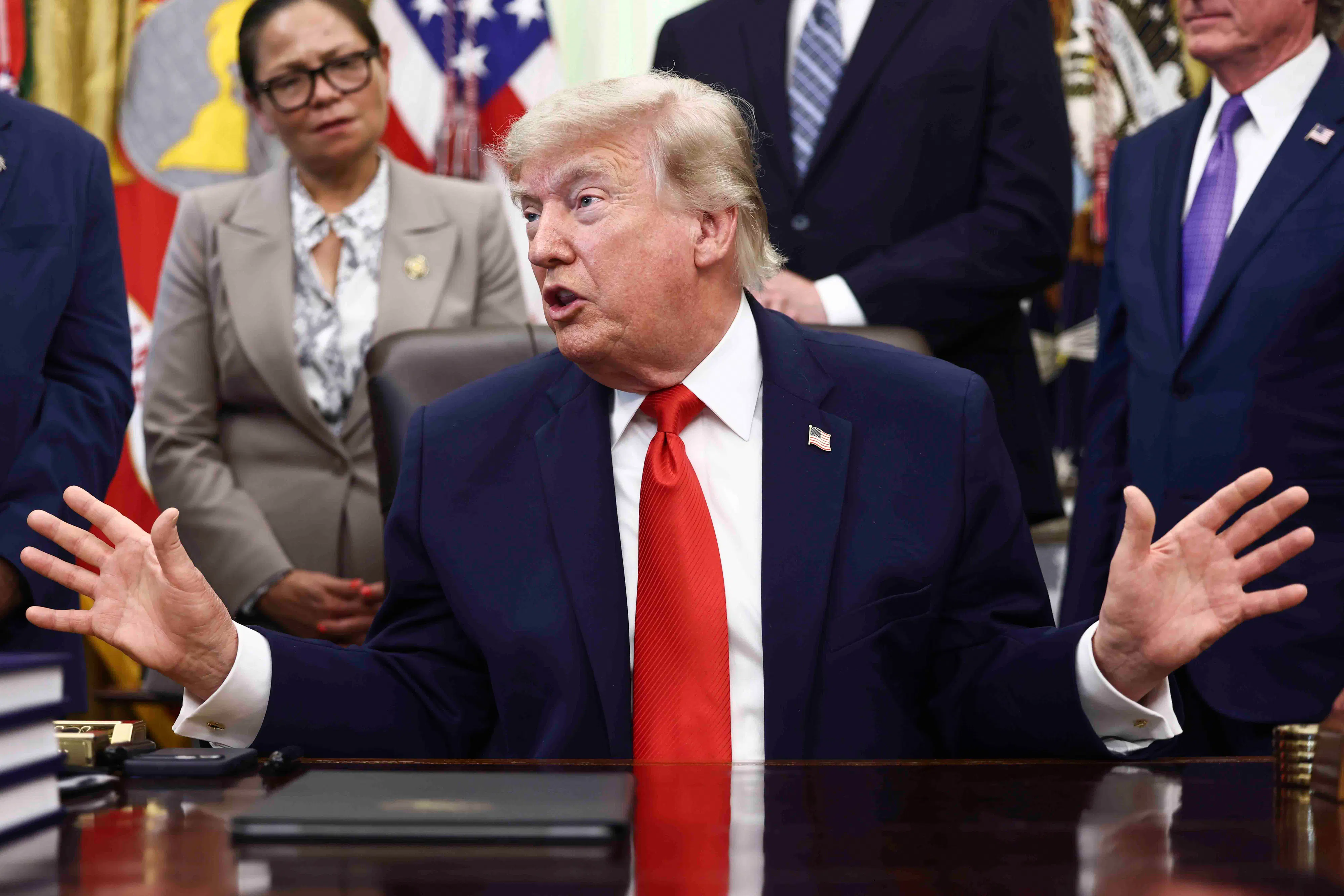Donald Trump sits at his desk in the Oval Office. He looks to the side and holds both hands out to the side while speaking.
