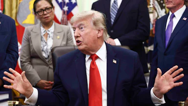 Donald Trump sits at his desk in the Oval Office. He looks to the side and holds both hands out to the side while speaking.