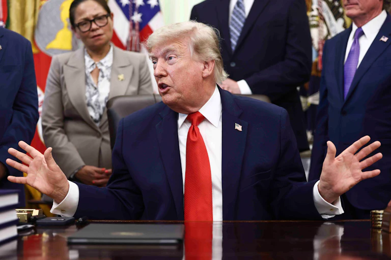 Donald Trump sits at his desk in the Oval Office. He looks to the side and holds both hands out to the side while speaking.