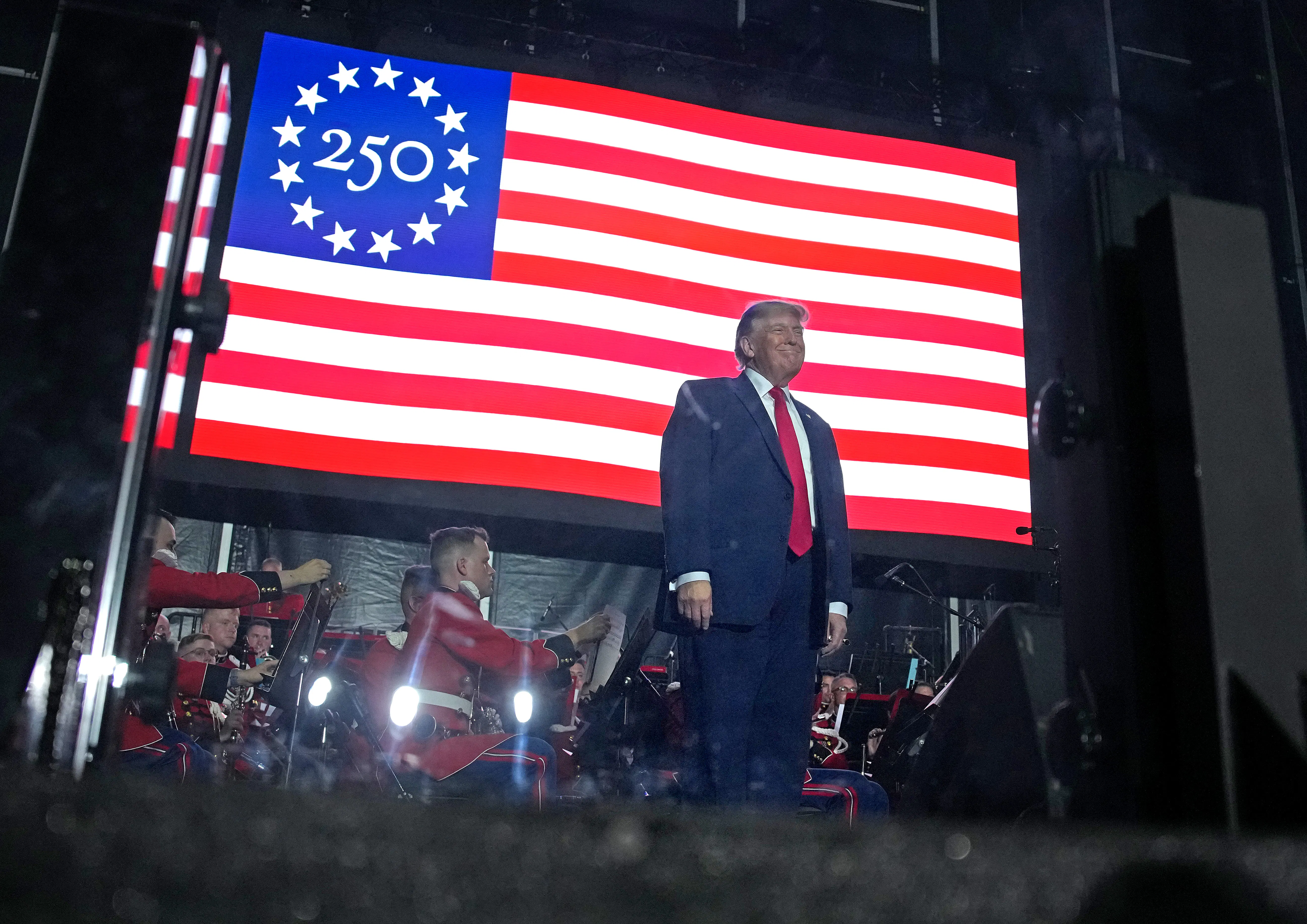 President Donald Trump stands on stage in front of a large U.S. flag with the number "250" as the stars.
