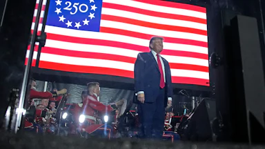 President Donald Trump stands on stage in front of a large U.S. flag with the number "250" as the stars.