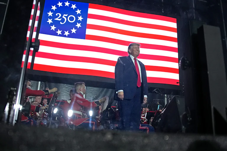 President Donald Trump stands on stage in front of a large U.S. flag with the number "250" as the stars.