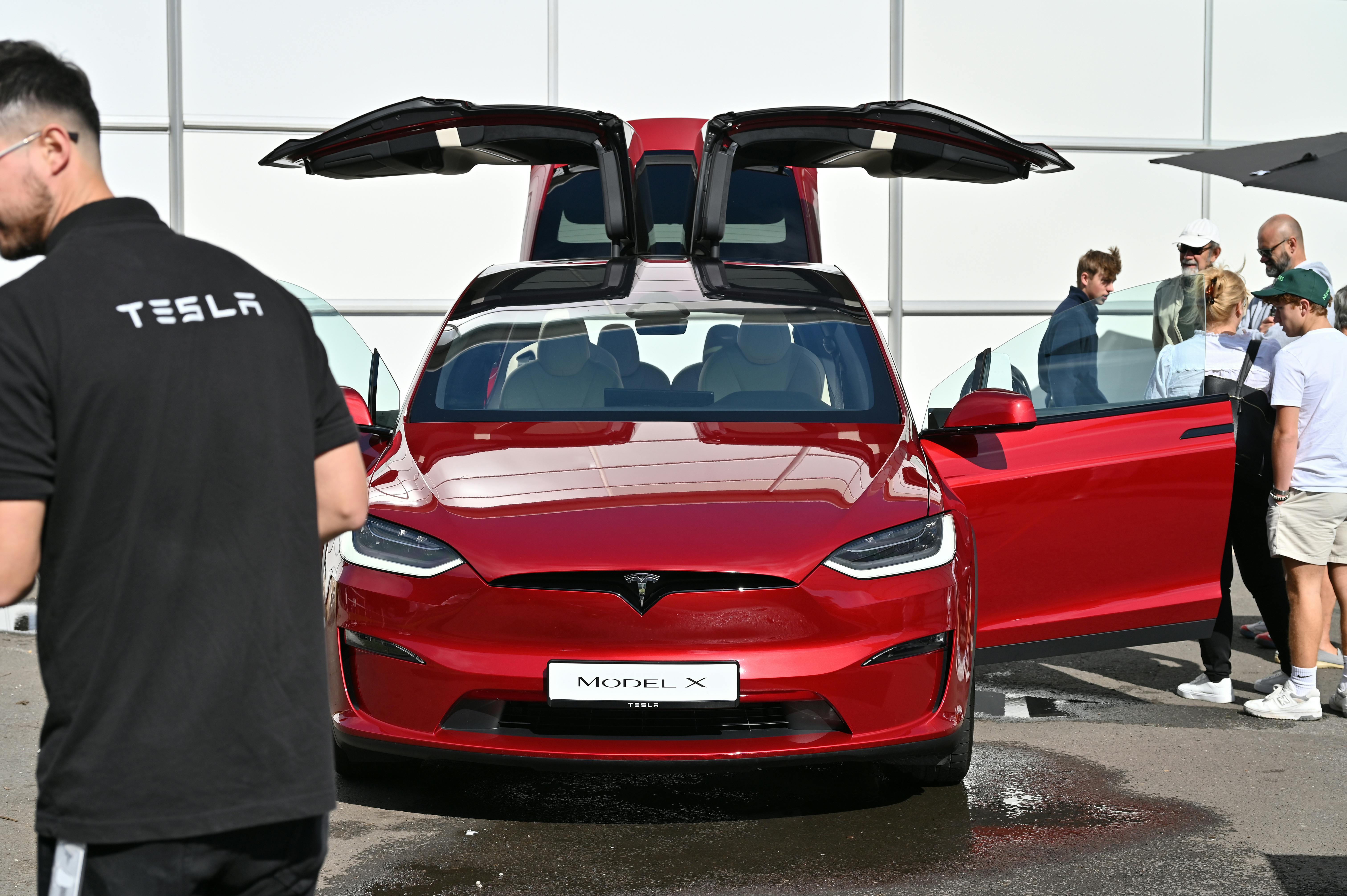 People cluster around a red Tesla Model X with its doors open.