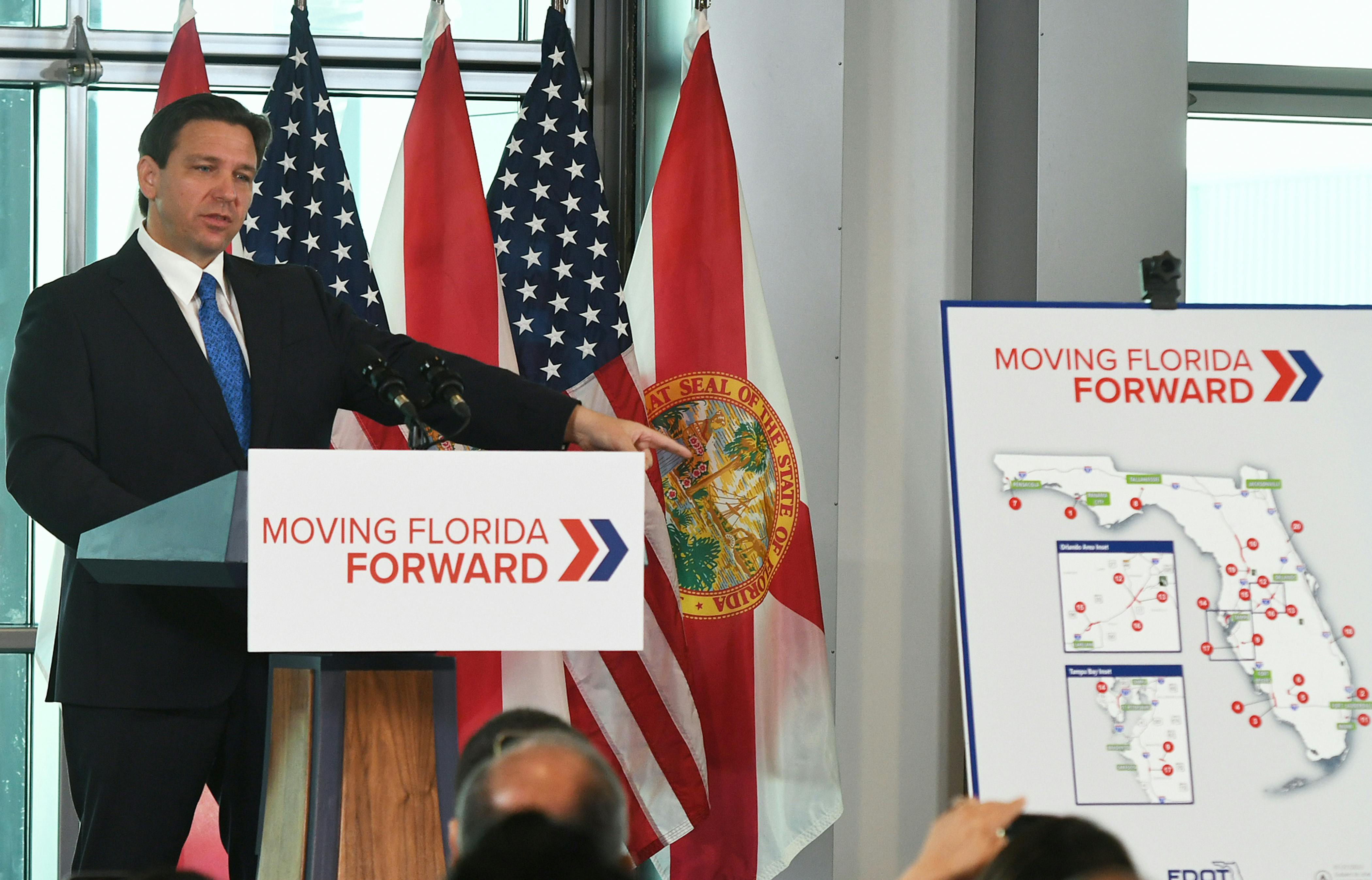 Ron DeSantis stands at a podium reading "Moving Florida Forward."