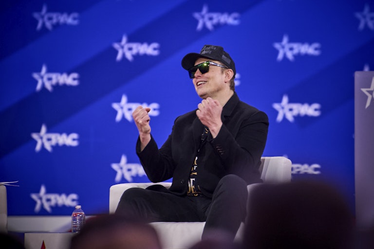 Elon Musk smiles and holds up his fists while seated onstage at CPAC