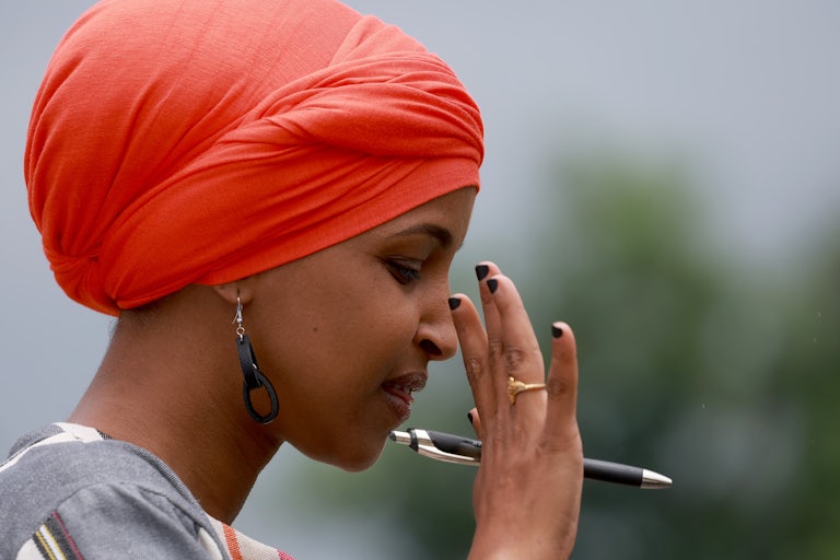 Ilhan Omar wears a bright red headwrap and puts her hand in the air as if tired