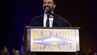 Zohran Mamdani smiles and stands at a podium that reads "New York Is Not For Sale."