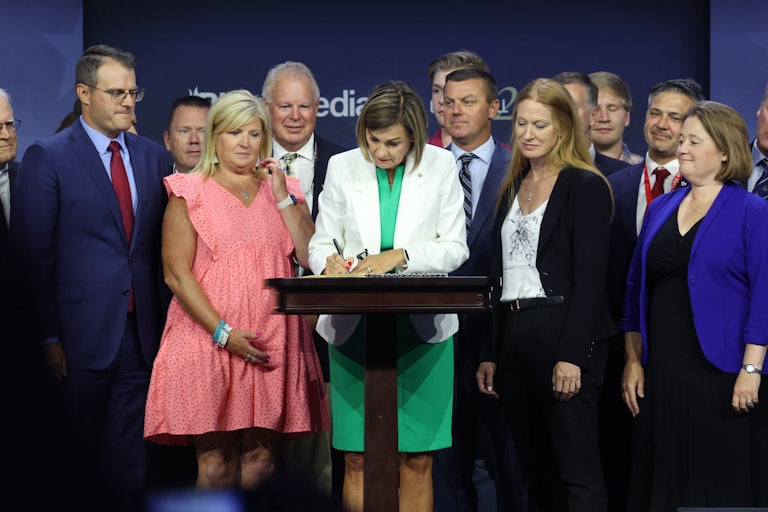 A woman wearing a white jacket and green dress signs a law on a lectern surrounded by white people.