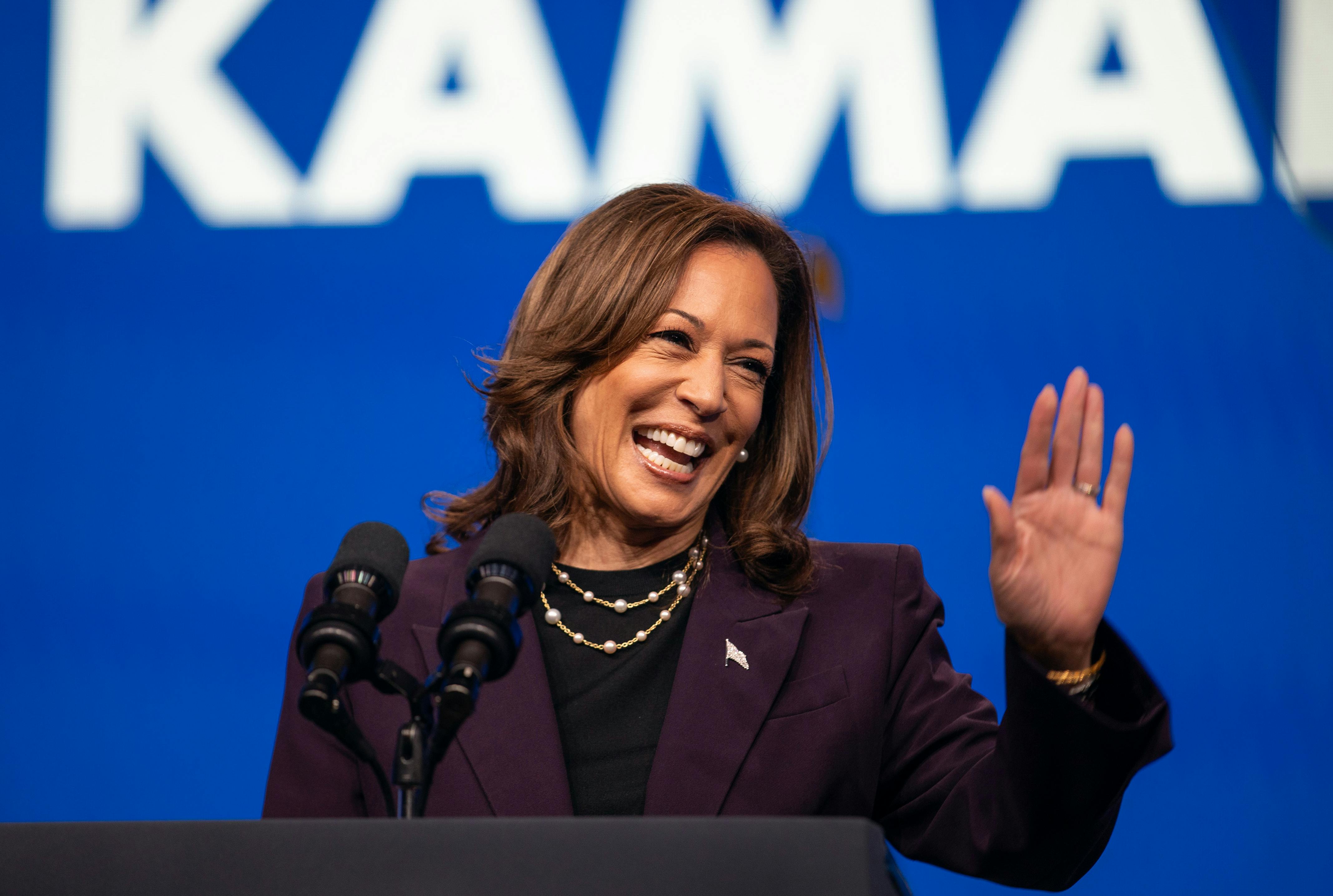 Kamala Harris, standing in front of a blue backdrop with white letters reading KAMALA, smiles broadly and holds up a single hand in a wave. 