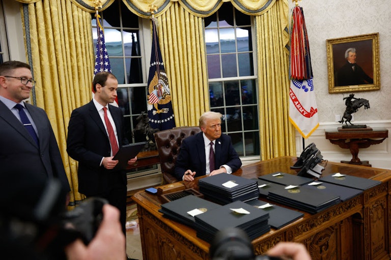 Trump sits at the presidential desk in the Oval Office with a stack of folders bearing executive orders in front of him, as two others look on while standing.