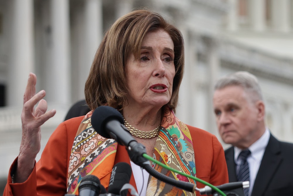 Nancy Pelosi speaks at a podium in front of the Capitol building while wearing an orange blazer and colorful scarf.