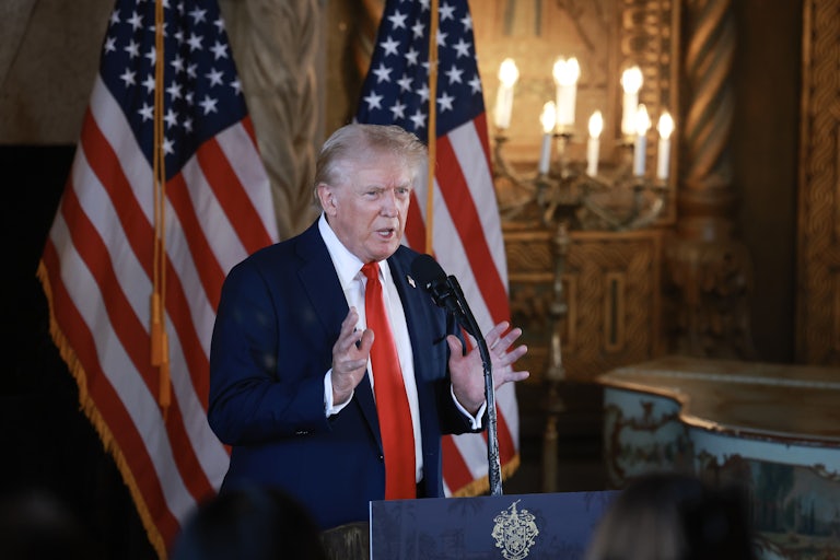 Donald Trump speaks animatedly at a lectern in Mar-a-Lago, brows furrowed and hands splayed. Two large U.S. flags stand behind him.