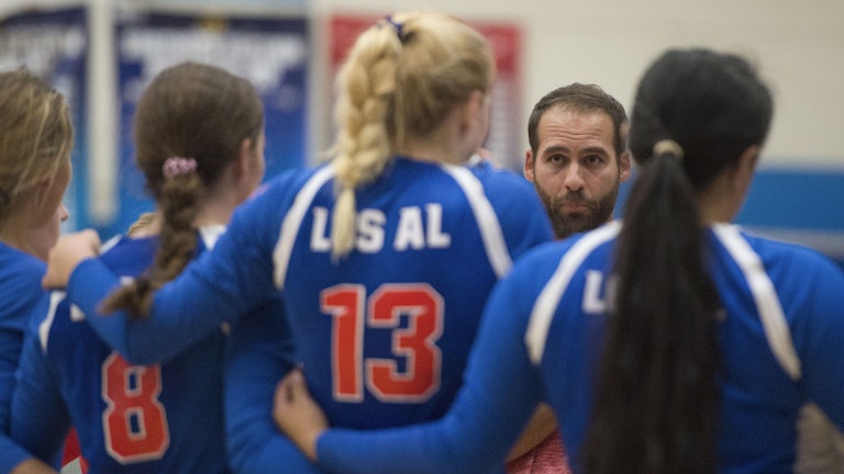 Four girls wearing volleyball jerseys link arms as their male coach looks on