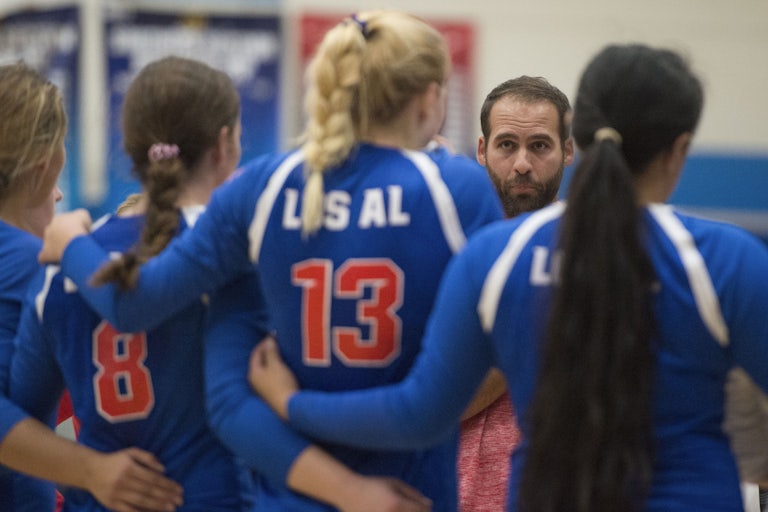 Four girls wearing volleyball jerseys link arms as their male coach looks on