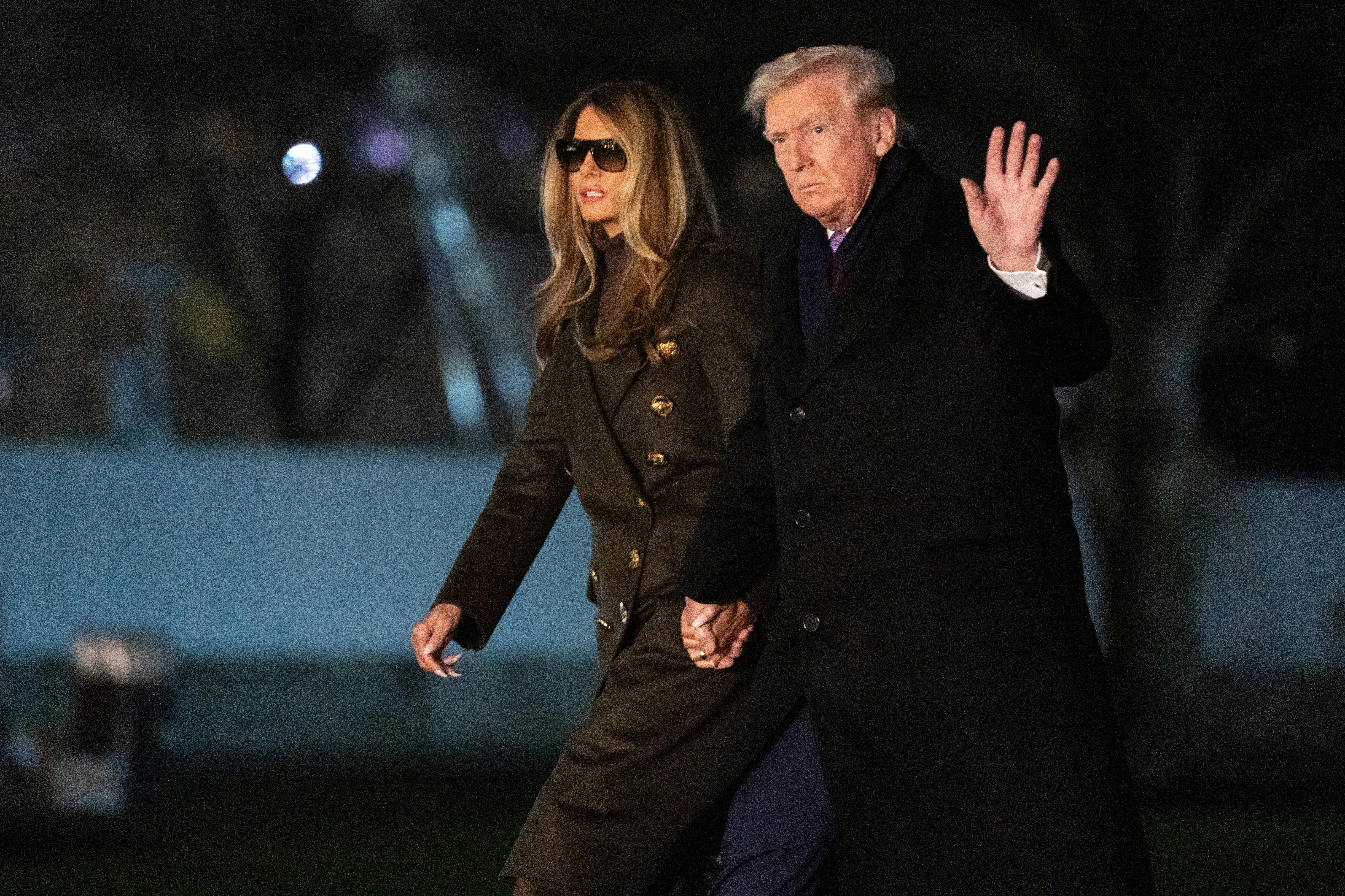 Donald and Melania Trump walk on the White House lawn hand in hand. Trump raises a hand for the camera.