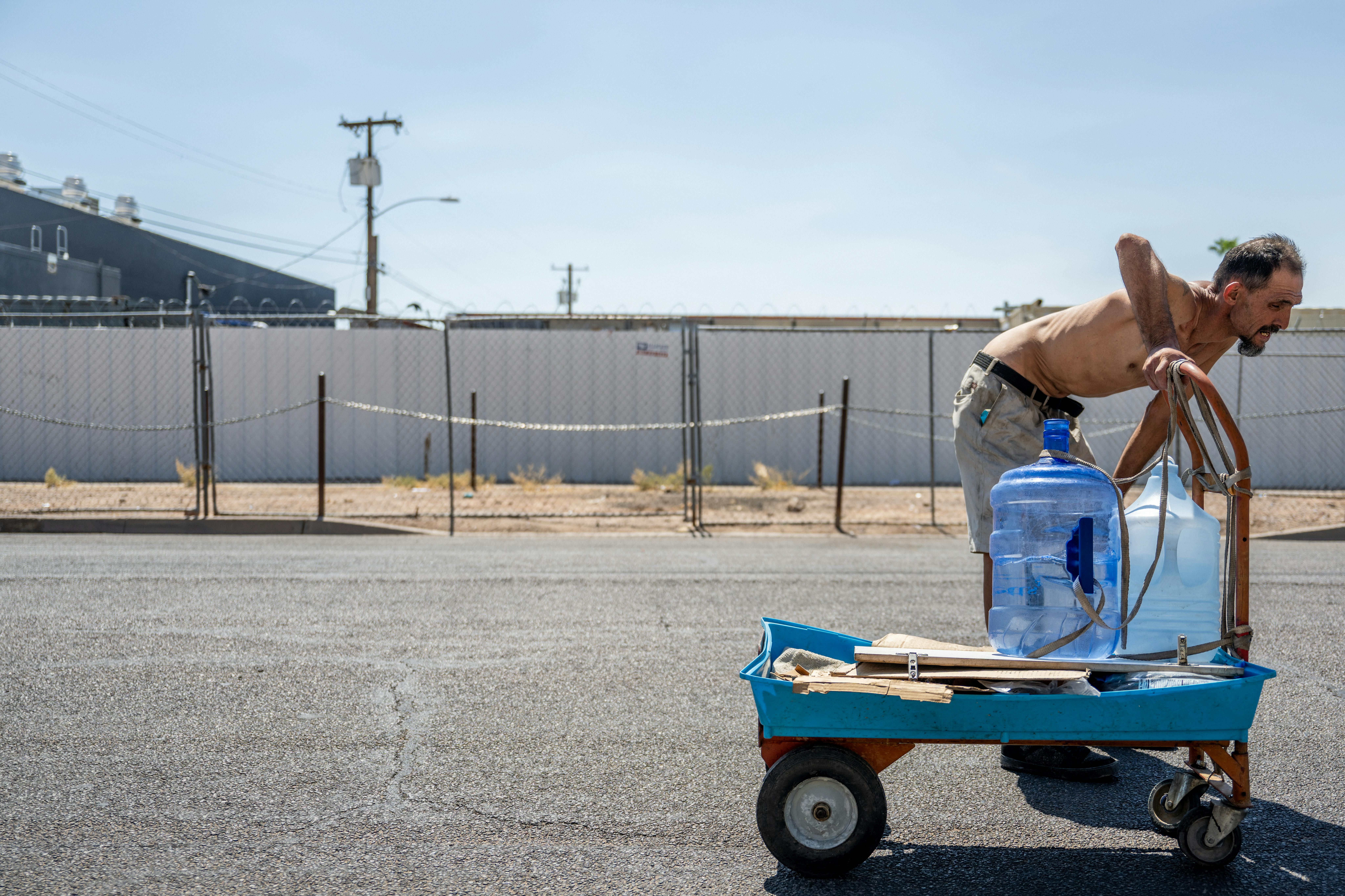 A person with no shirt rests their hands on their knees in the middle of a bright street while pushing a cart with a jug of water on top.