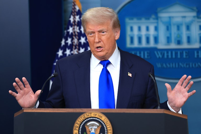 Donald Trump raises his hands as if in defense and speaks at the podium in the White House press briefing room.