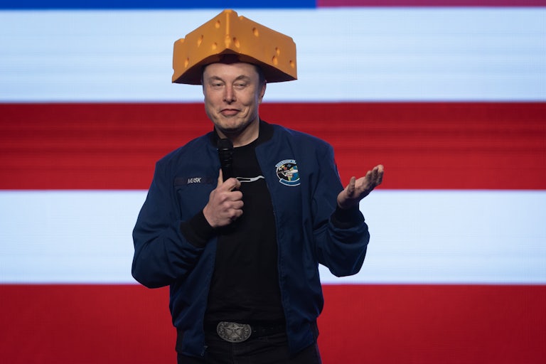 Elon Musk wears a hat that looks like a block of speech and holds a microphone and gestures while onstage at a rally in Wisconsin.