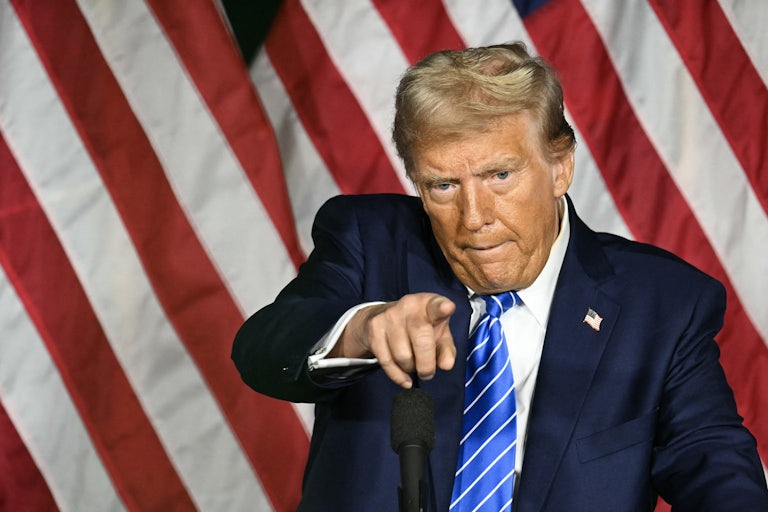 Donald Trump points at something or someone off stage. U.S. flags are behind him.