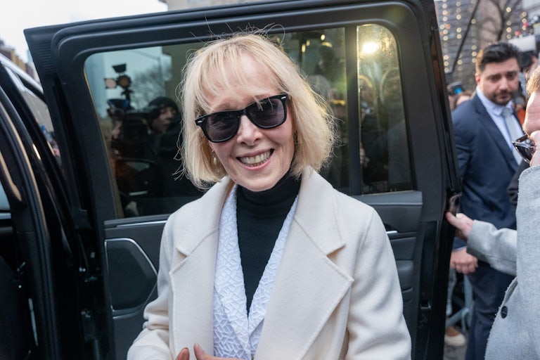 E. Jean Carroll smiles wearing a beige pea coat, a white blazer, a black turtleneck, and sunglasses. A car door is open behind her, and there are other people and cameras in the background.