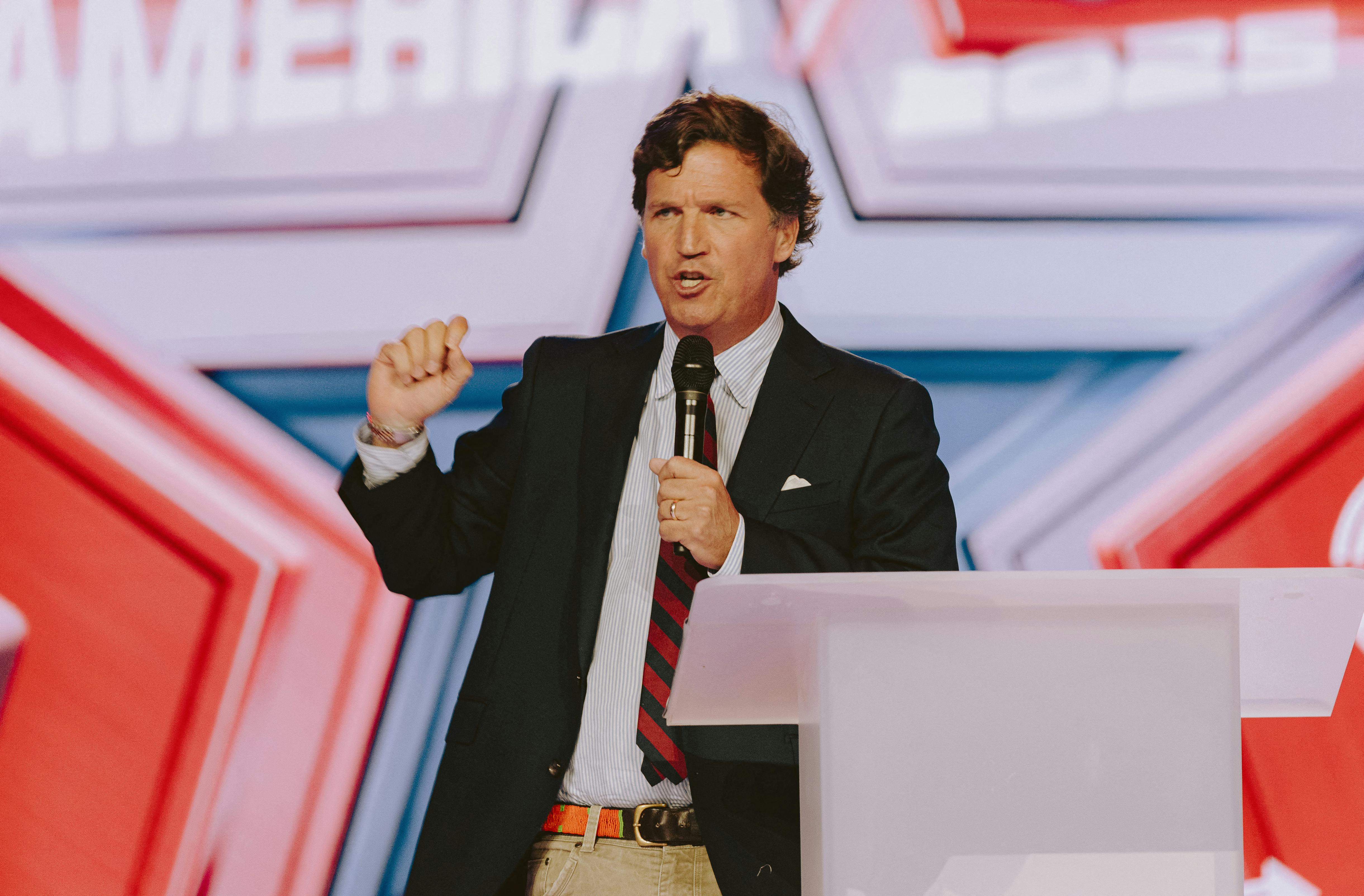Tucker Carlson speaks at a podium while holding a handheld mic.