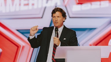 Tucker Carlson speaks at a podium while holding a handheld mic.