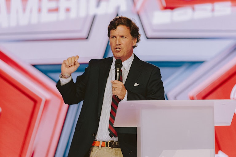 Tucker Carlson speaks at a podium while holding a handheld mic.