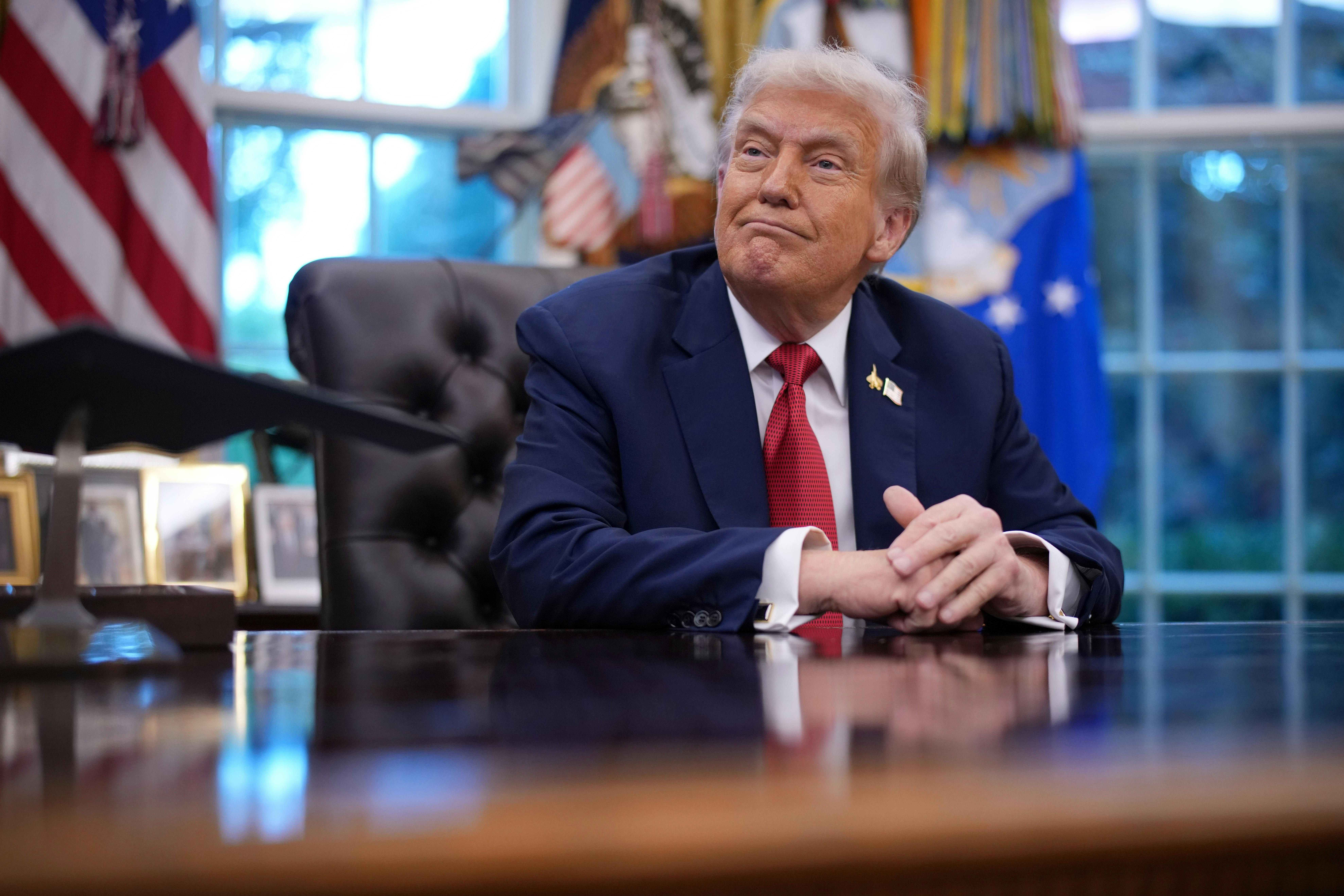 Donald Trump looks as someone hands him a folder while he's seated behind his desk in the Oval Office of the White House.