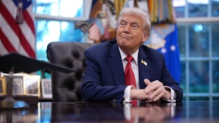 Donald Trump looks as someone hands him a folder while he's seated behind his desk in the Oval Office of the White House.