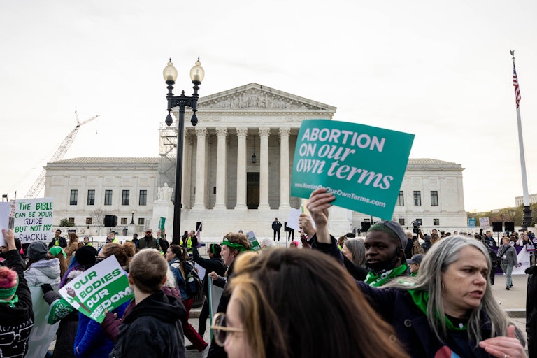 Protesters gather in front of the Supreme Court. One woman with gray hair holds a sign that reads "Abortion on our own terms"