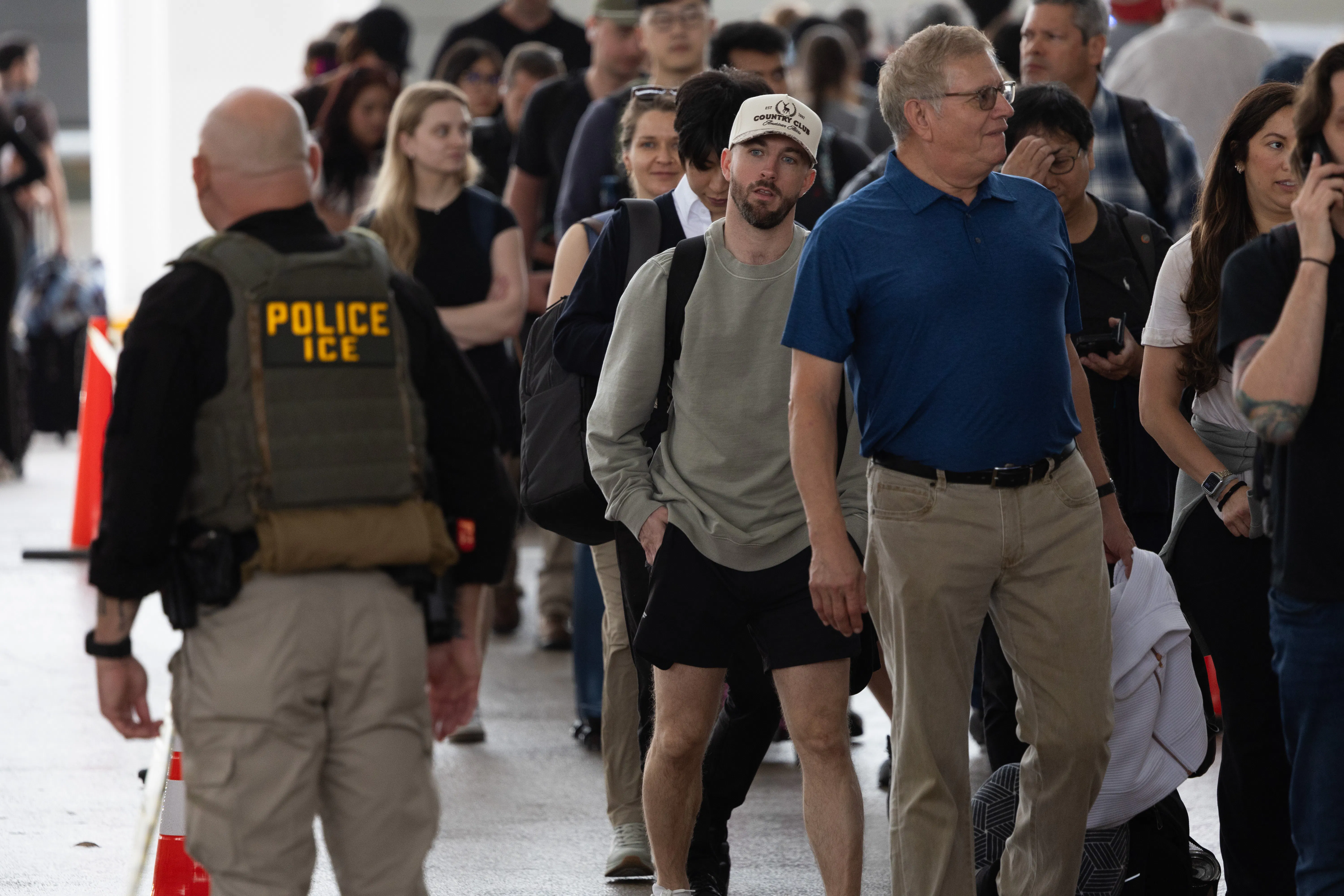 An ICE agent passes by travelers waiting in line at the airport.