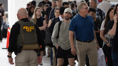 An ICE agent passes by travelers waiting in line at the airport.