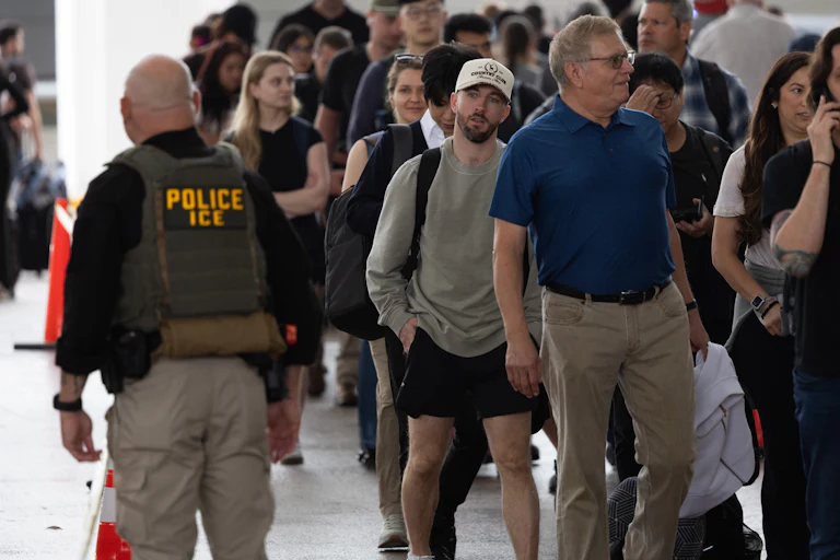 An ICE agent passes by travelers waiting in line at the airport.