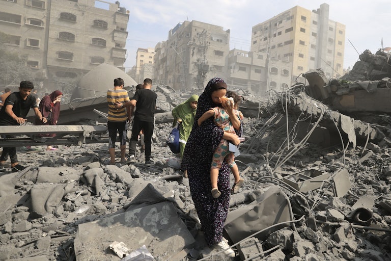 A woman cradles a child as she stands with others amidst rubble.