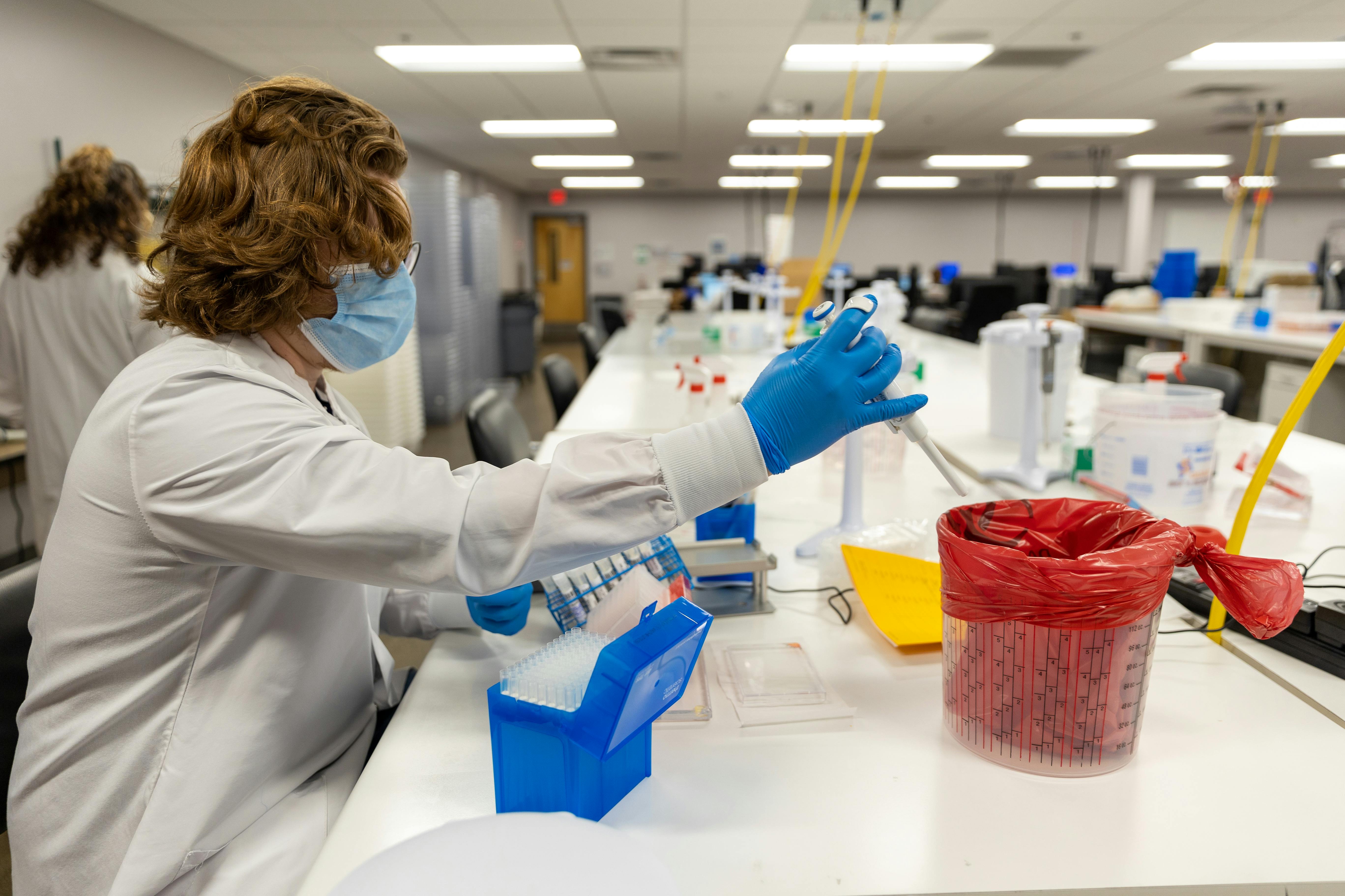 A person in a lab coat works with pipettes while seated.
