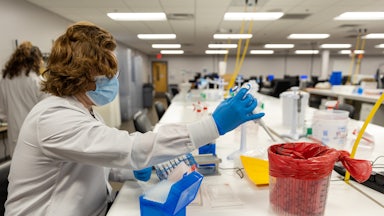 A person in a lab coat works with pipettes while seated.