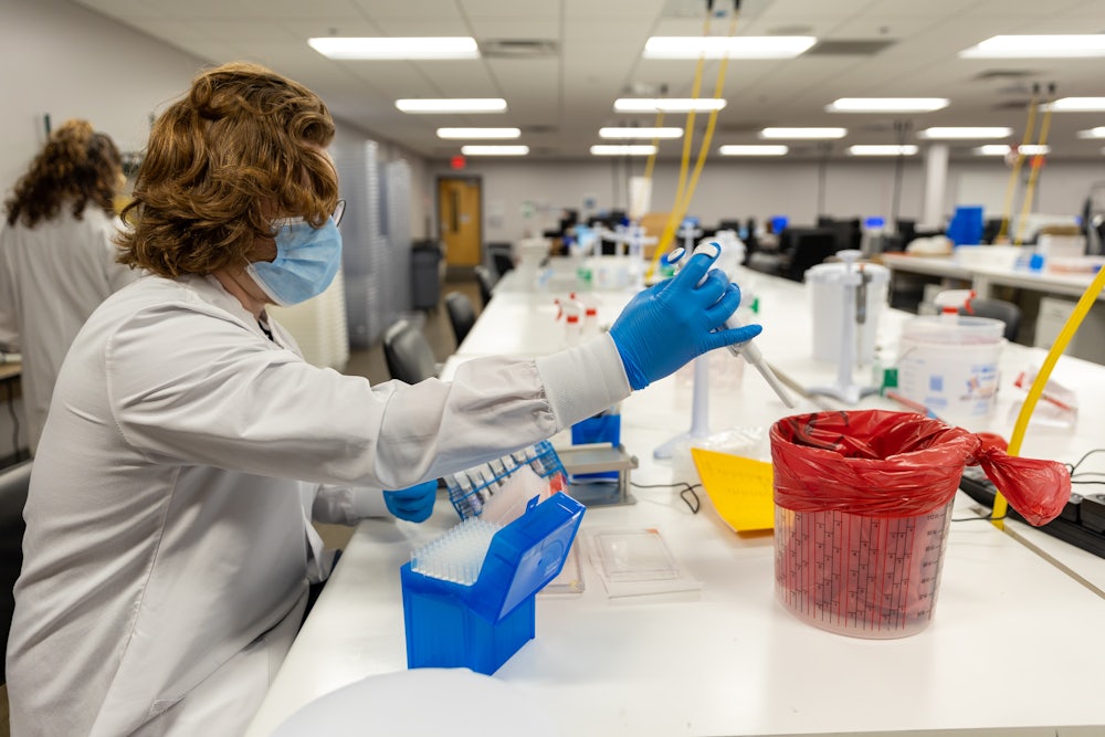 A person in a lab coat works with pipettes while seated.