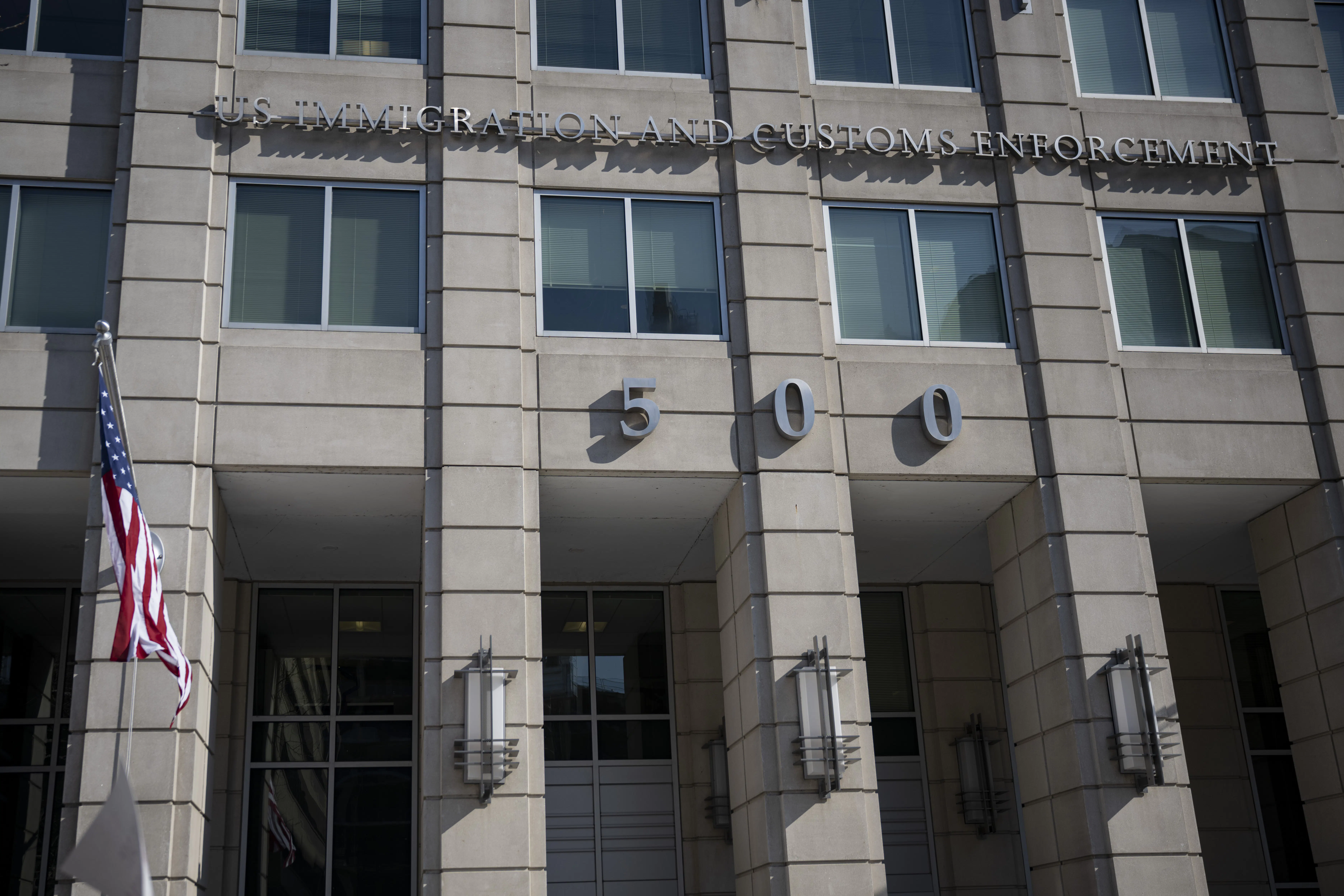 The ICE headquarters building, reading IMMIGRATION AND CUSTOMS ENFORCEMENT above the number 500 across the building's concrete front, with a flagpole on the side.