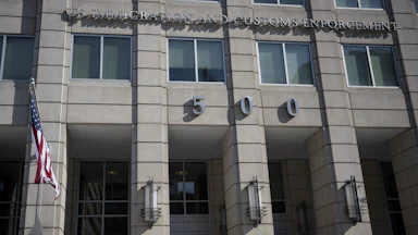 The ICE headquarters building, reading IMMIGRATION AND CUSTOMS ENFORCEMENT above the number 500 across the building's concrete front, with a flagpole on the side.